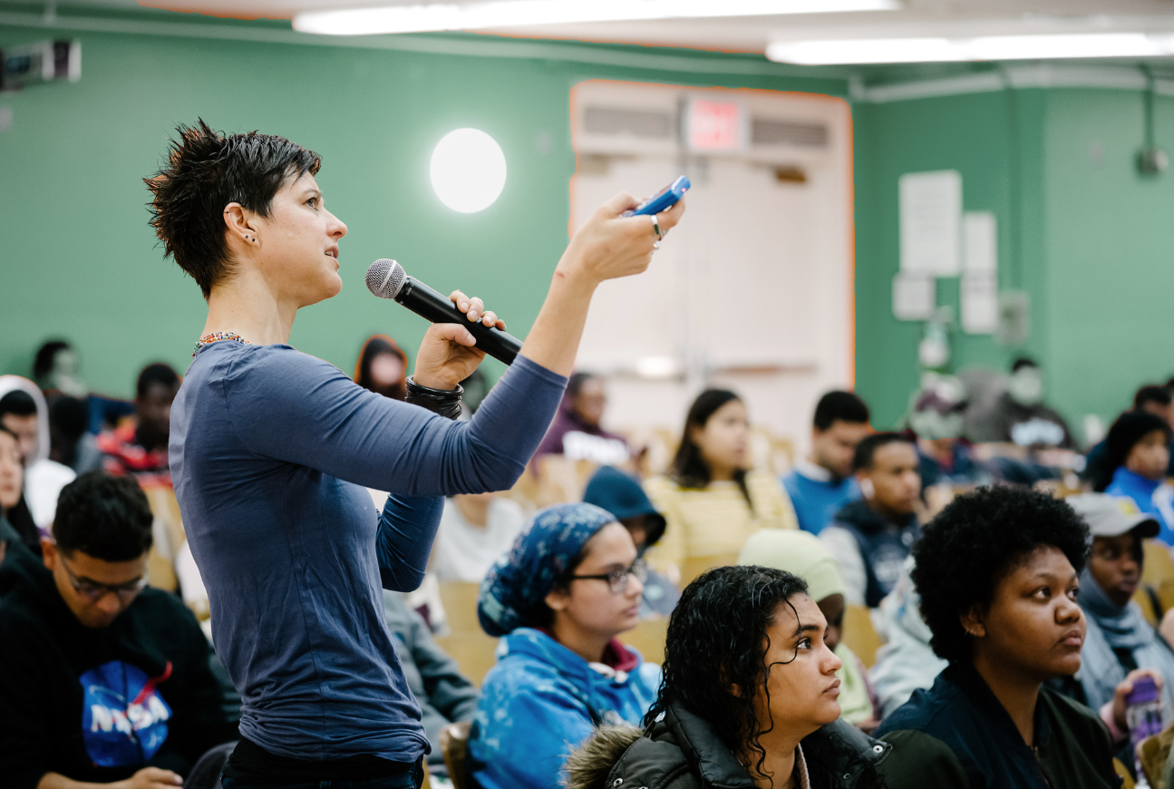 A woman speaking into a microphone, looking sideways, a remote control in her hand, teaching to a group of students seated in a classroom.