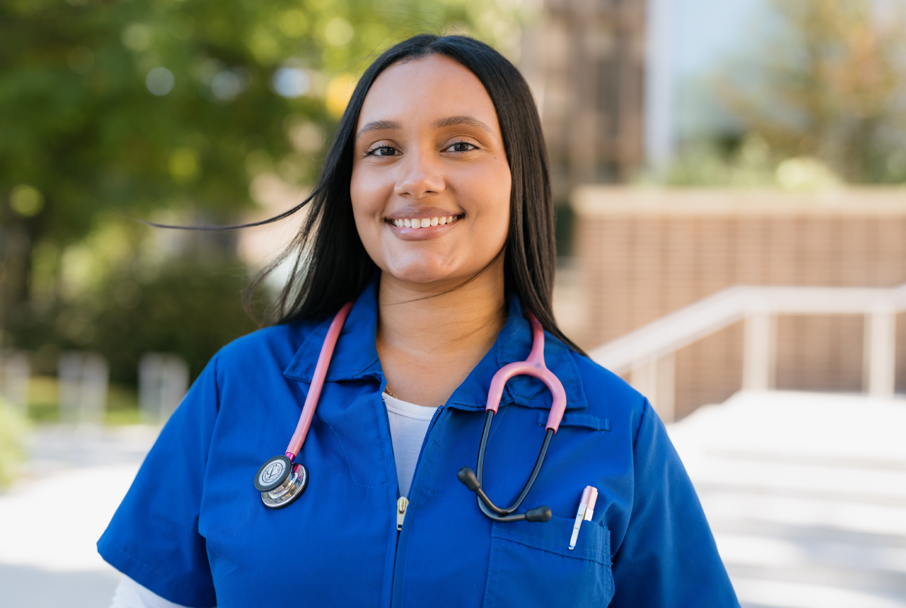 Smiling healthcare worker wearing royal blue scrubs and a pink stethoscope.