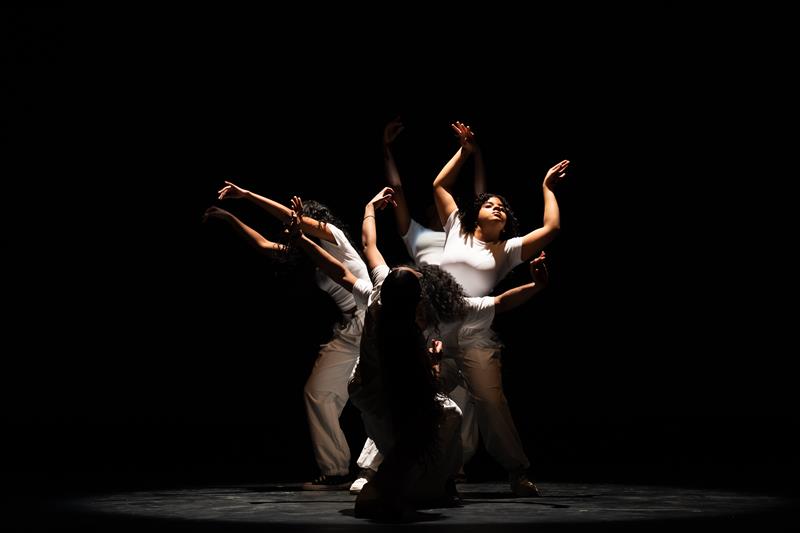 Group of dancers in white t-shirts and beige pants close together with their arms stretched out in different directions.
