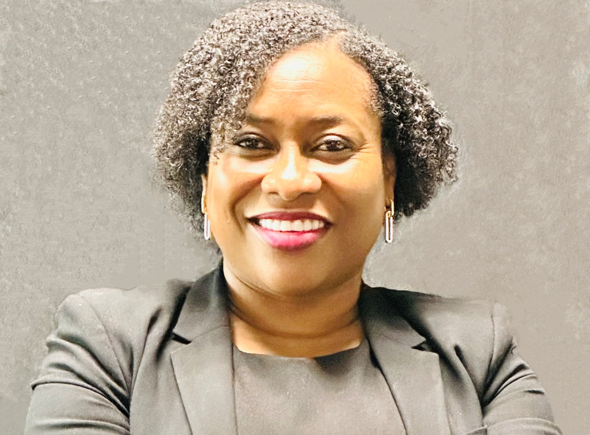 AFRICAN AMERICAN FEMAL WITH SHORT CURLY HAIR, WEARING A BLACK BLAZER AND BLOUSE, GOLD COLORED EARNINGS, AND RED LIPSTICK. STANDING IN FRONT OF A BLACK BACKGROUND