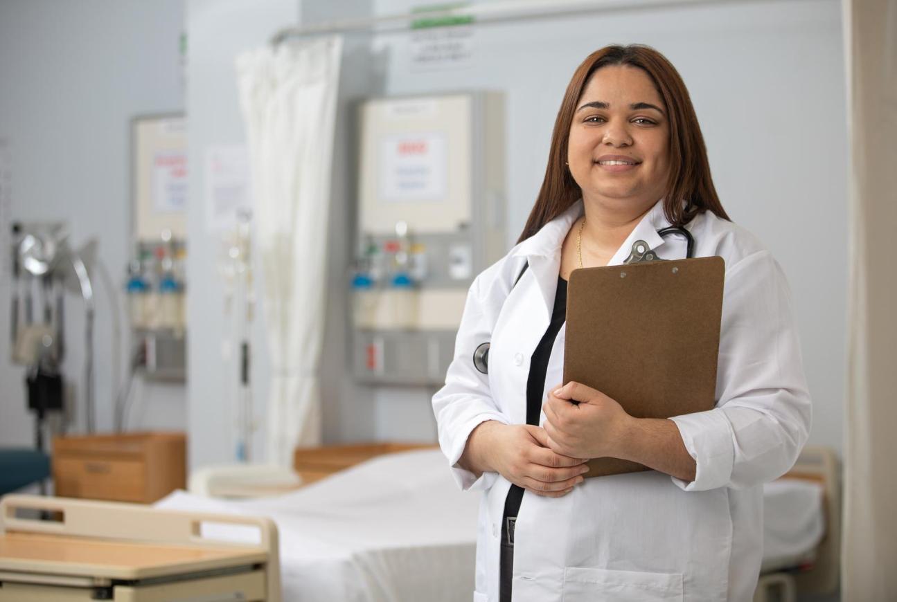 Healthcare professional in a lab coat holding a clipboard in a clinical training room.