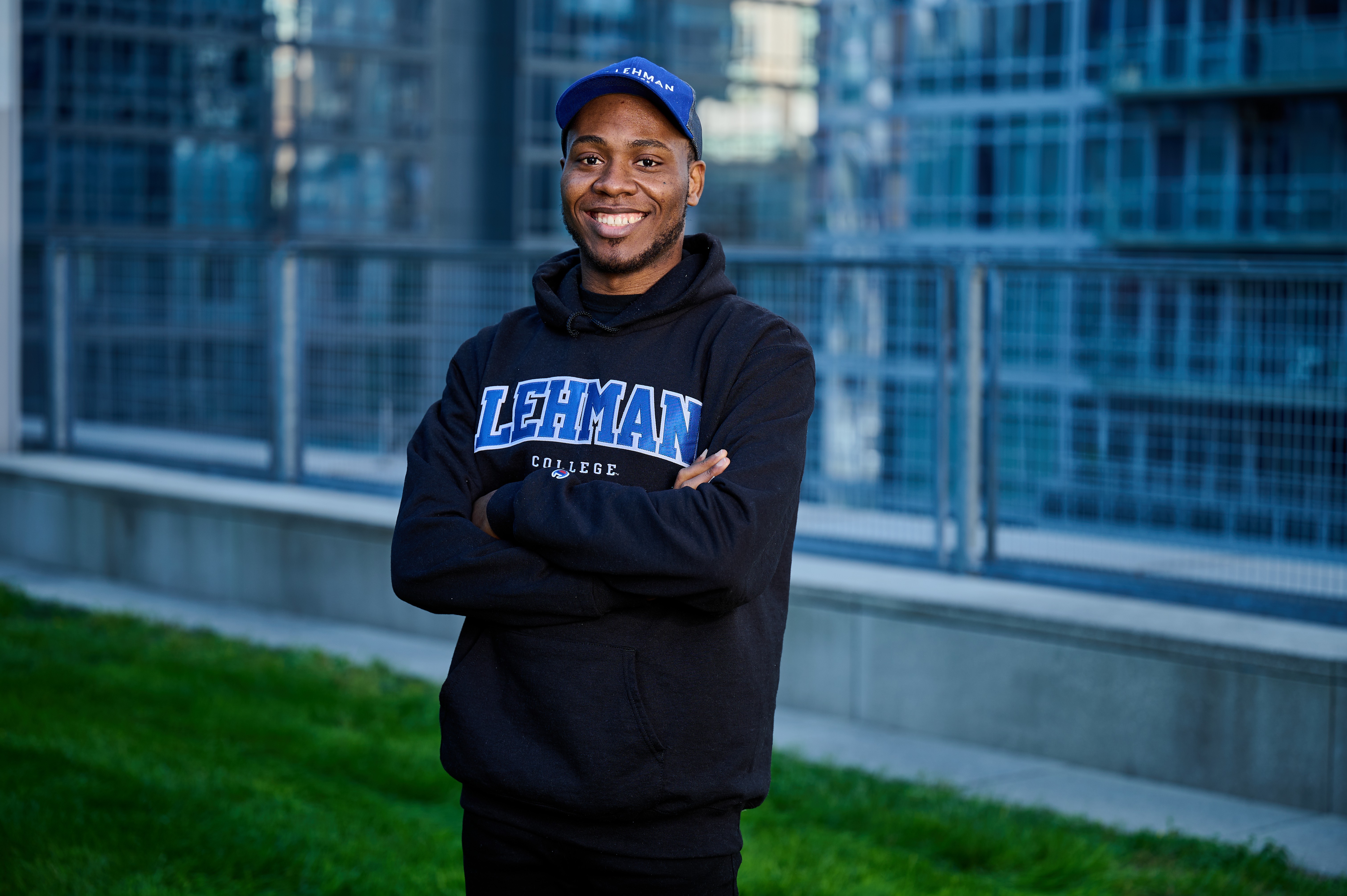 A student posing in front of a building. He is wearing a blue baseball cap and a Lehman College sweatshirt.