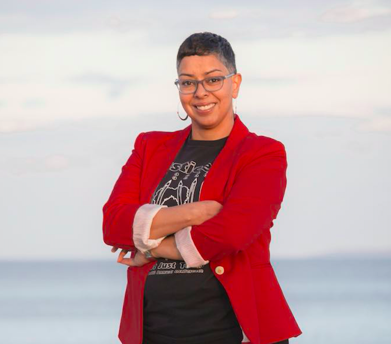 Woman with short hair and glasses in red jacket standing with arms folded.