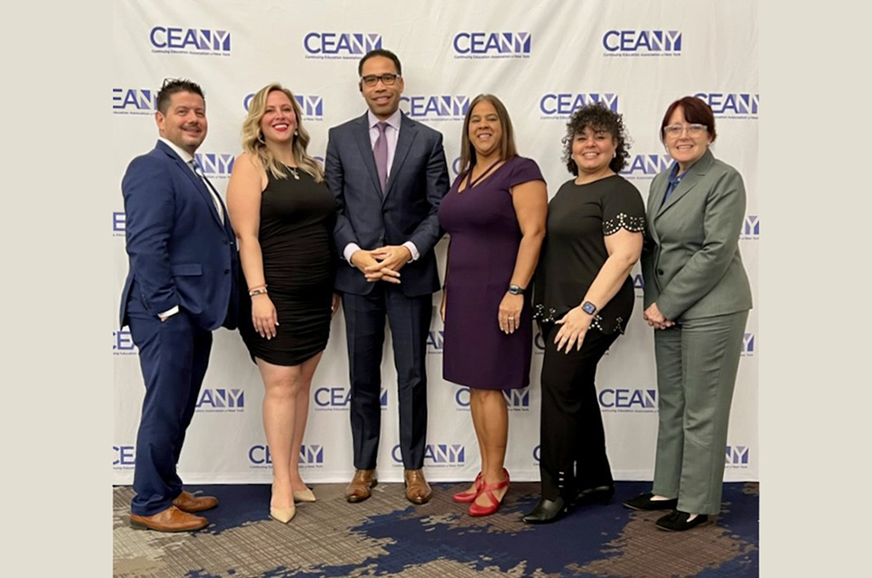 A group of six people, men and women, posing together in front of a white backdrop with blue logos reading CEANY printed on it.