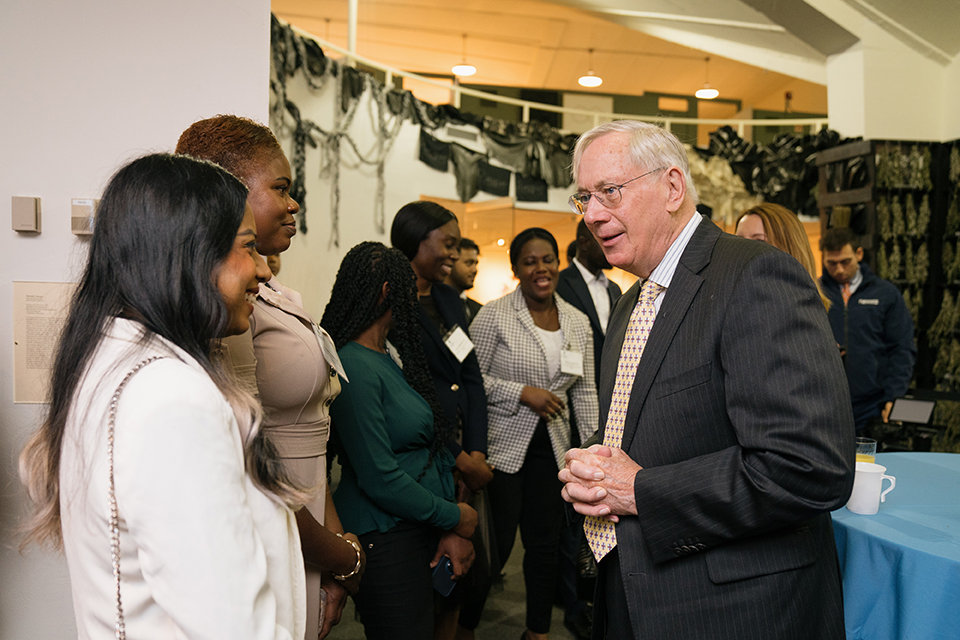 A grey-haired white man wearing a dark suit speaking with two women of color at a reception..