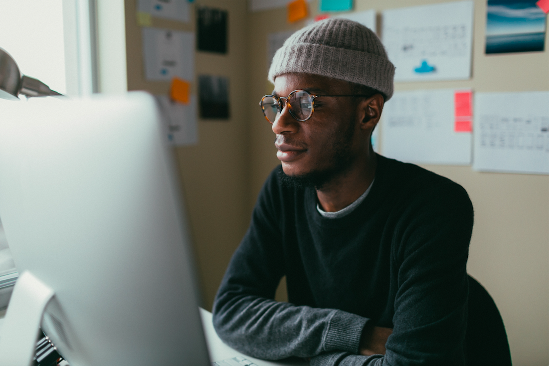 Man wearing glasses and beanie hat sitting at a computer.