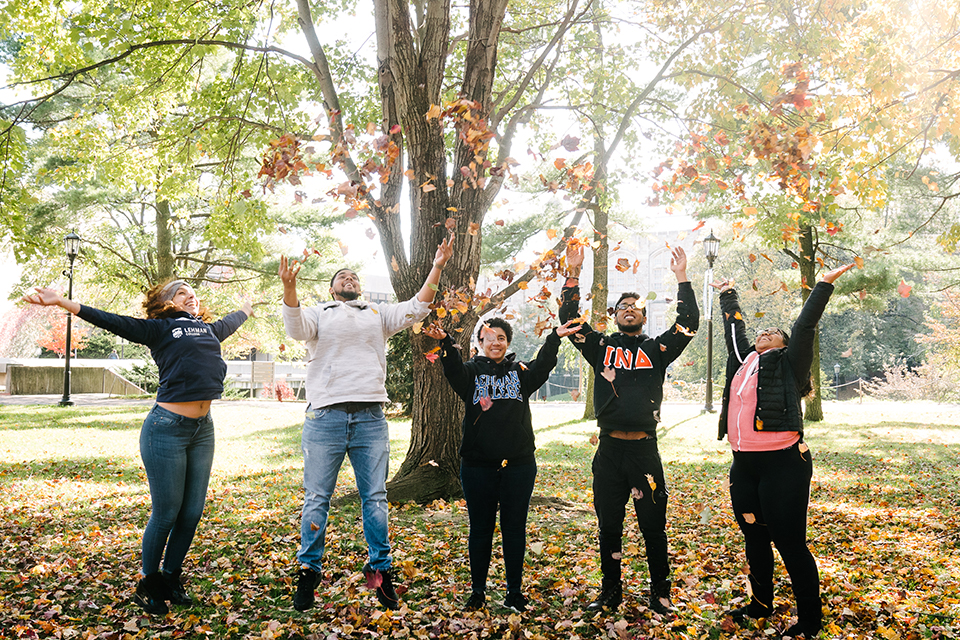 Five people throwing fallen leaves into the air. They are standing under trees.