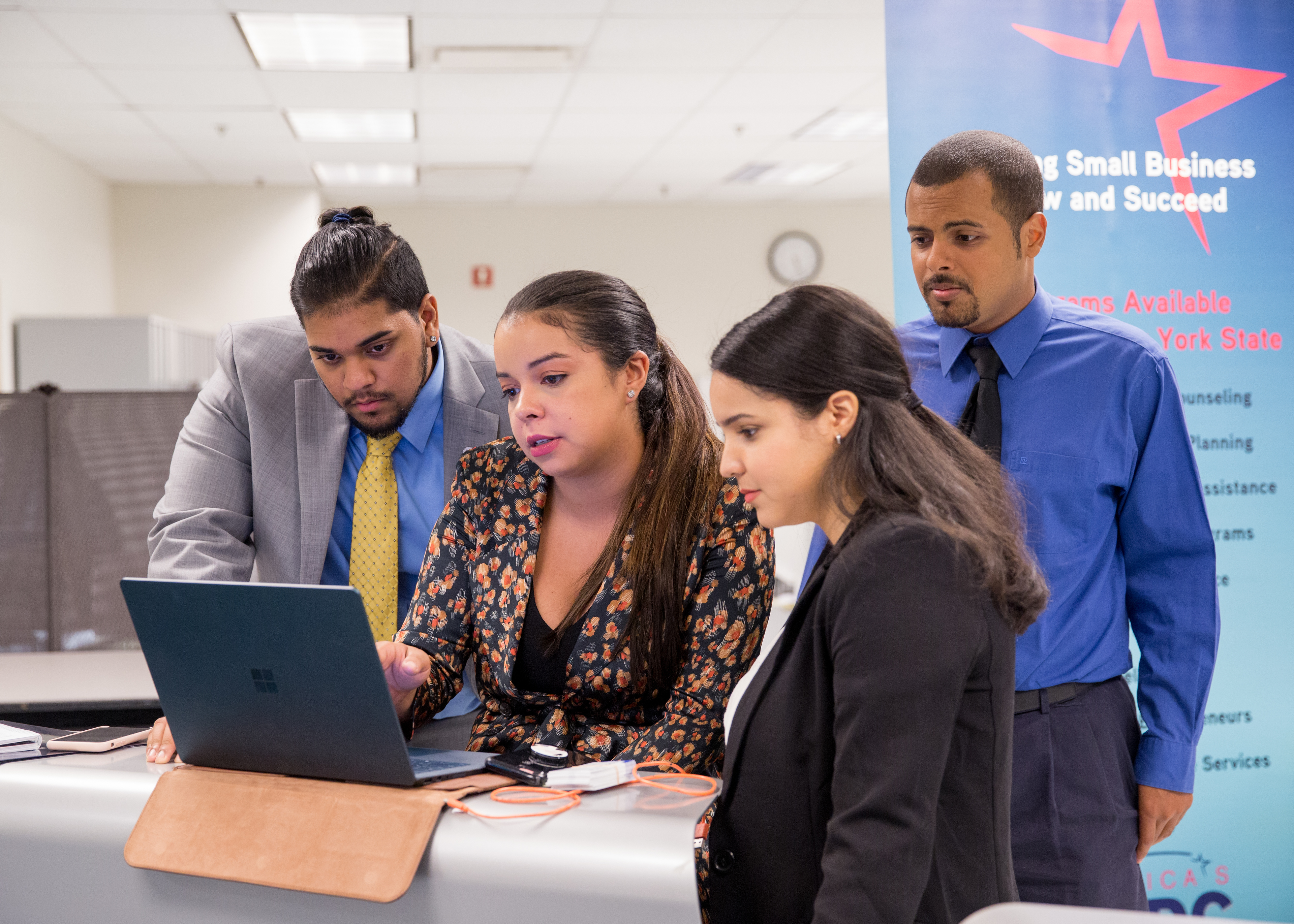 A standing group of young people, two men and two women,  in an office looking at a computer