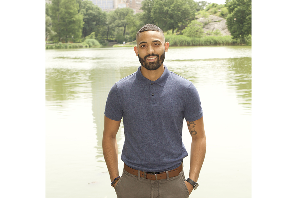 Hispanic man with close-cropped hair in a dark shirt and brown pants posing in front of a lake with trees in the background..