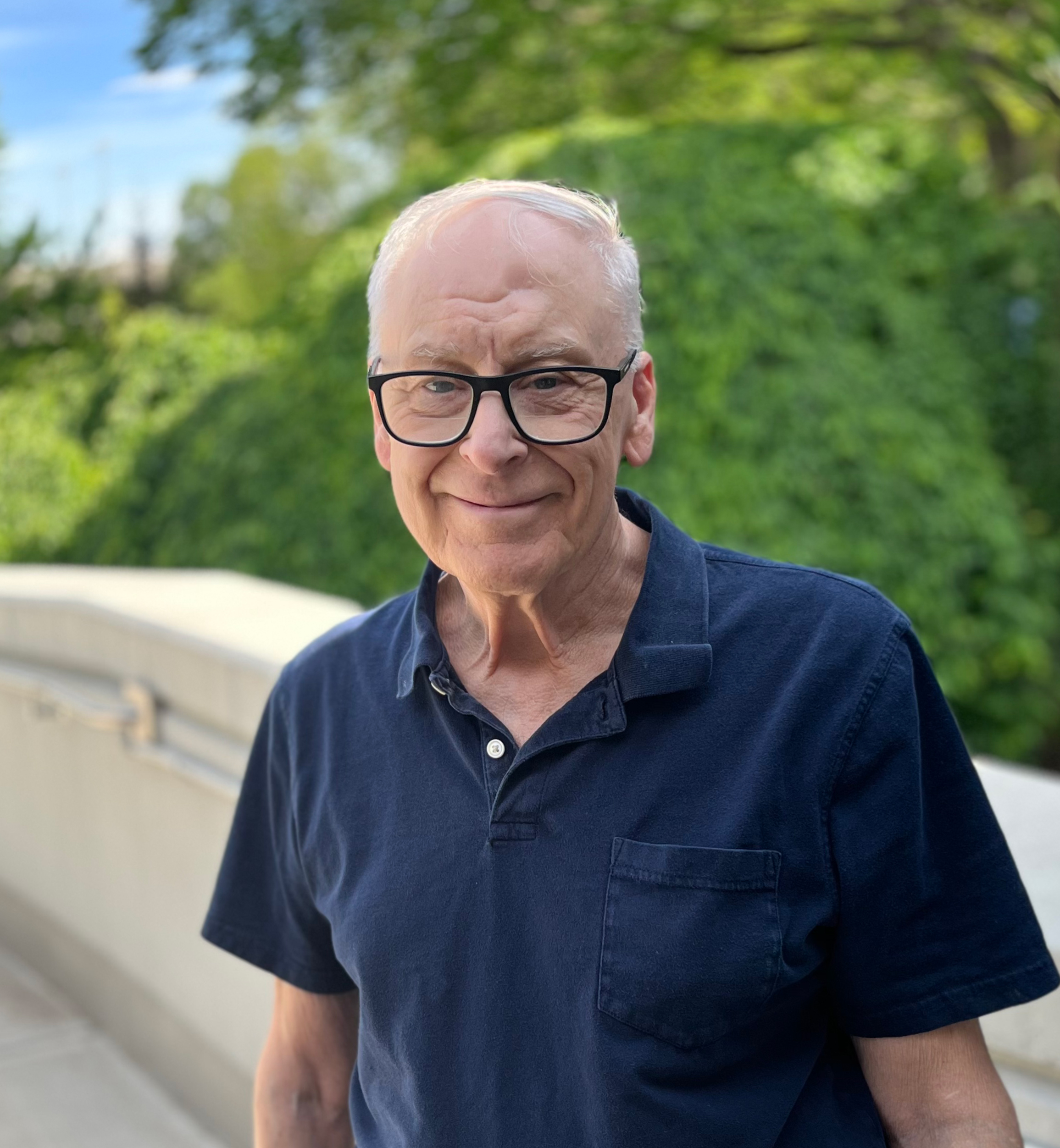 An older man with white hair, glasses, and a blue shirt poses with trees in the background.