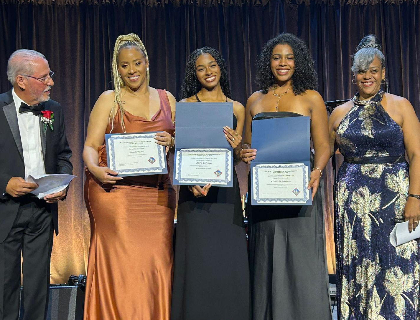 Three women in formal gowns holding certificates, with a man and a woman on either side of them.